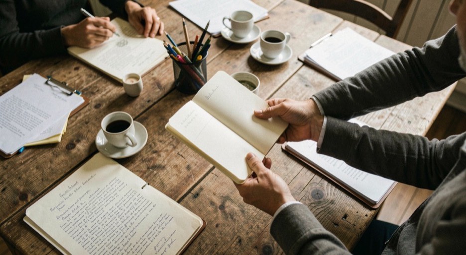 Deux mains échangent un carnet de notes au-dessus d’une table claire, une fiche imprimée glissée entre les pages