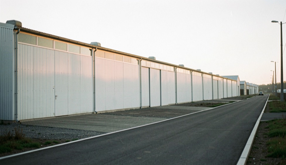 Vue d’ensemble d’une zone artisanale au lever du jour, hangars de tôle claire alignés le long d’une voirie neuve
